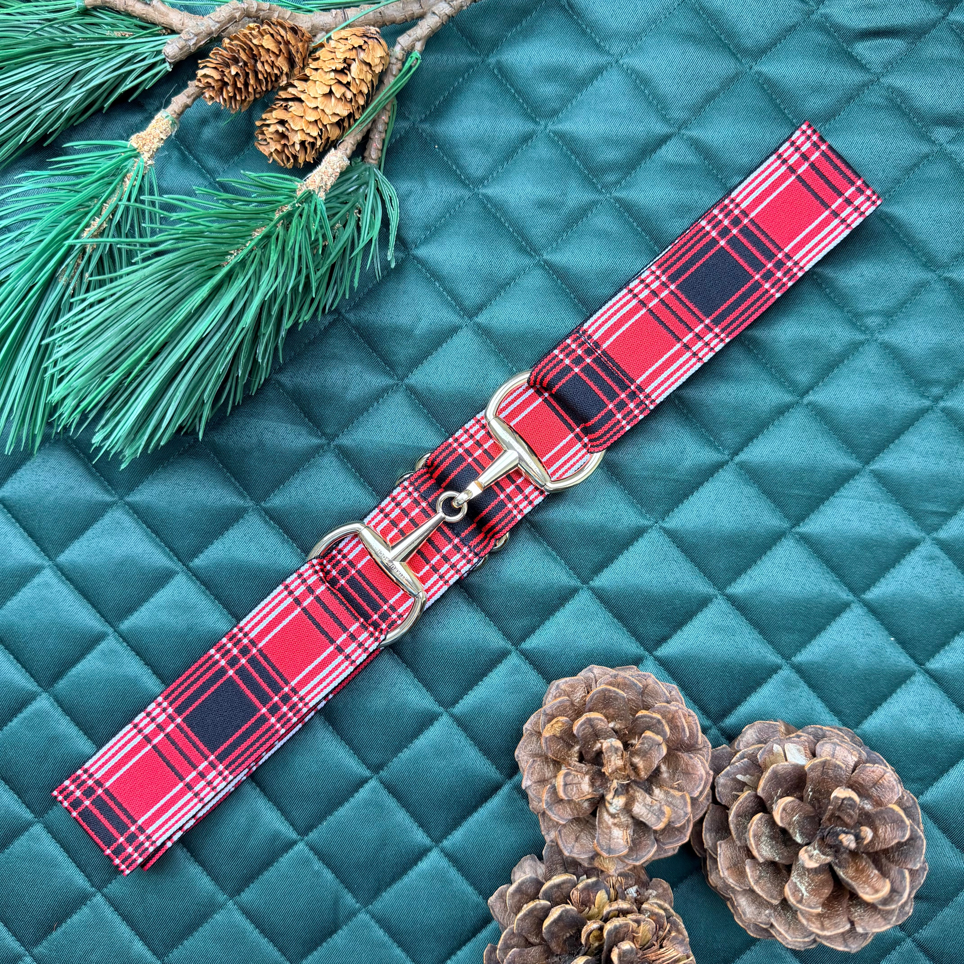 Red plaid equestrian belt with a gold snaffle bit buckle on a green saddle pad with pine branches and cones.