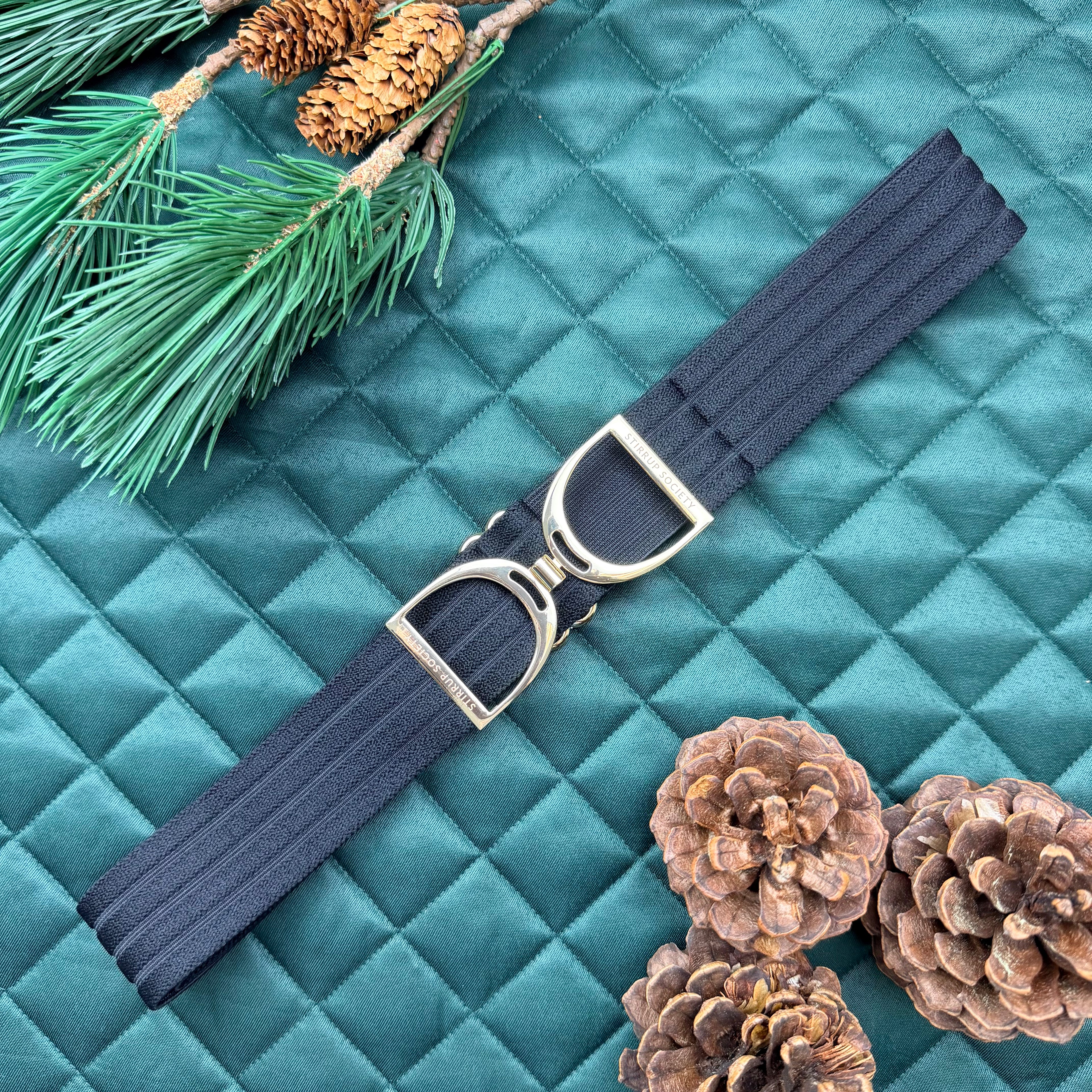 Black equestrian belt with buckle on a green saddle pad with pine cones and branches.
