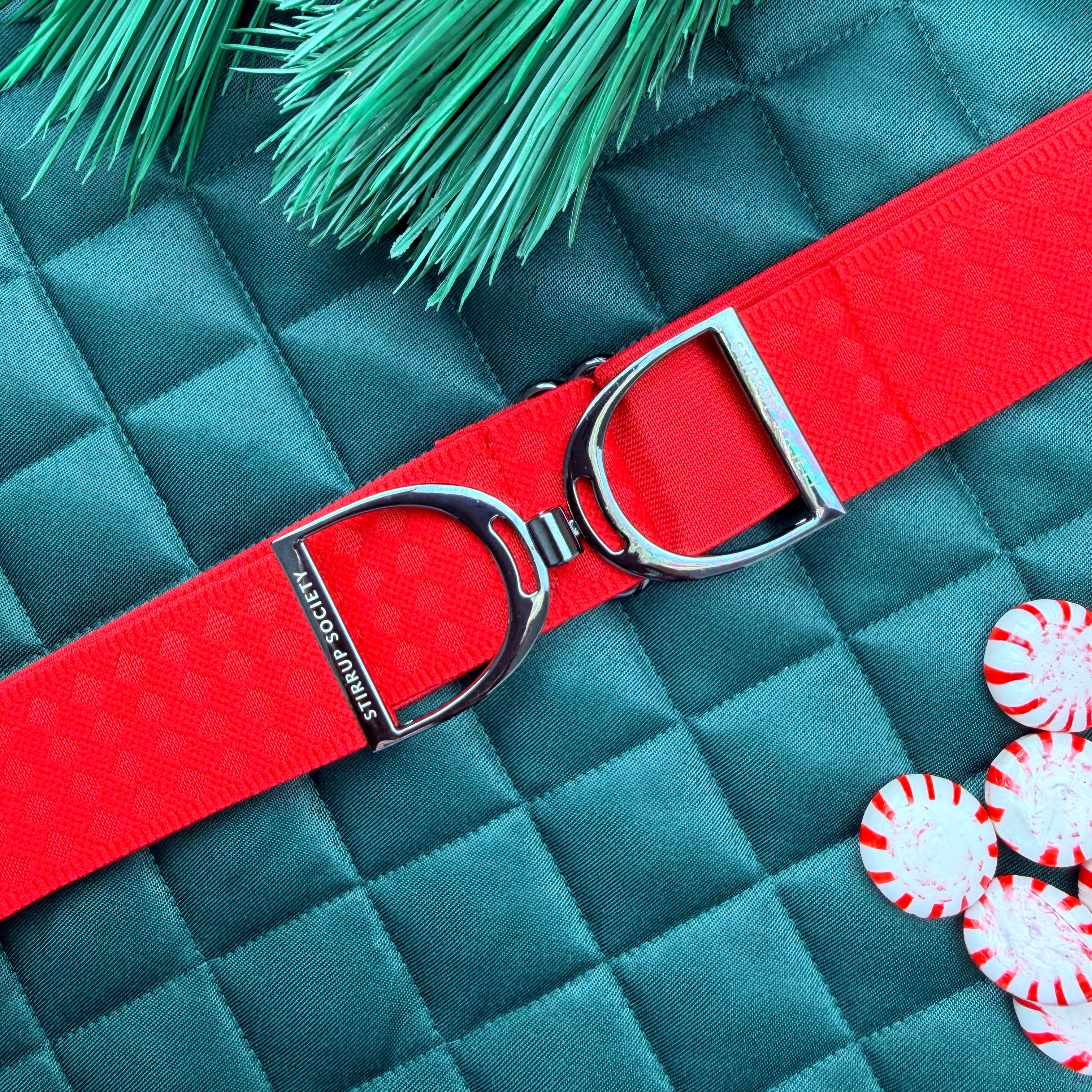 Red equestrian belt with silver stirrup buckle on a green saddle pad peppermint candies and green leaves.
